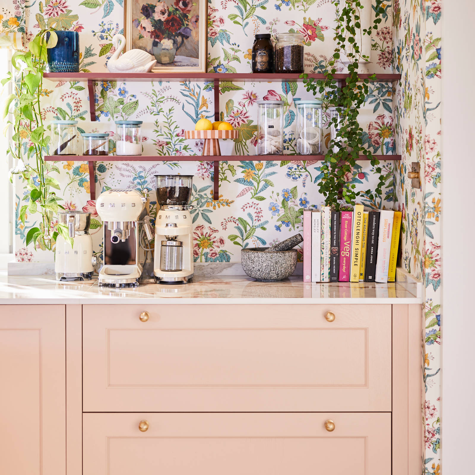 Coffee area in Sophie Robinson's maximalist kitchen, with shaker style Ludlow Chalk blush cabinetry, open shelving on a wall with a floral wallpaper and Smeg coffee machines on countertop.