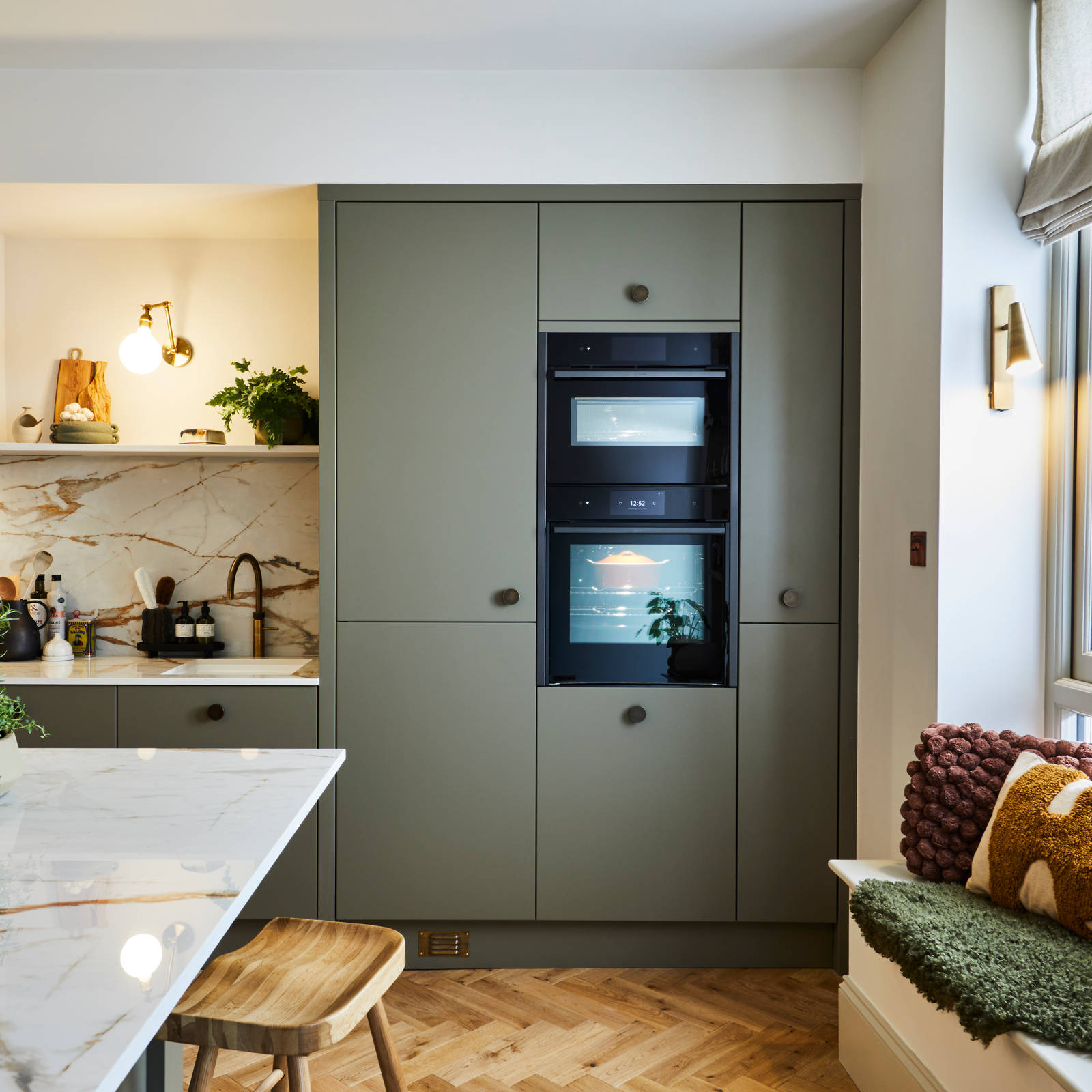 Built-in ovens in tall slab Soho Balmoral cabinets, a bench with earth tone pillows and a kitchen island with a wooden barstool and a plant