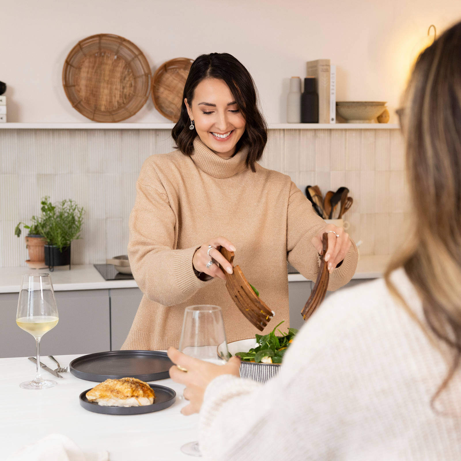 Yasmin Wyatt plating salad from a lage bowl on the kitchen island with plates and glasses of wine as someone is sitting by the opposite end of the kitchen island with their back to the camera.