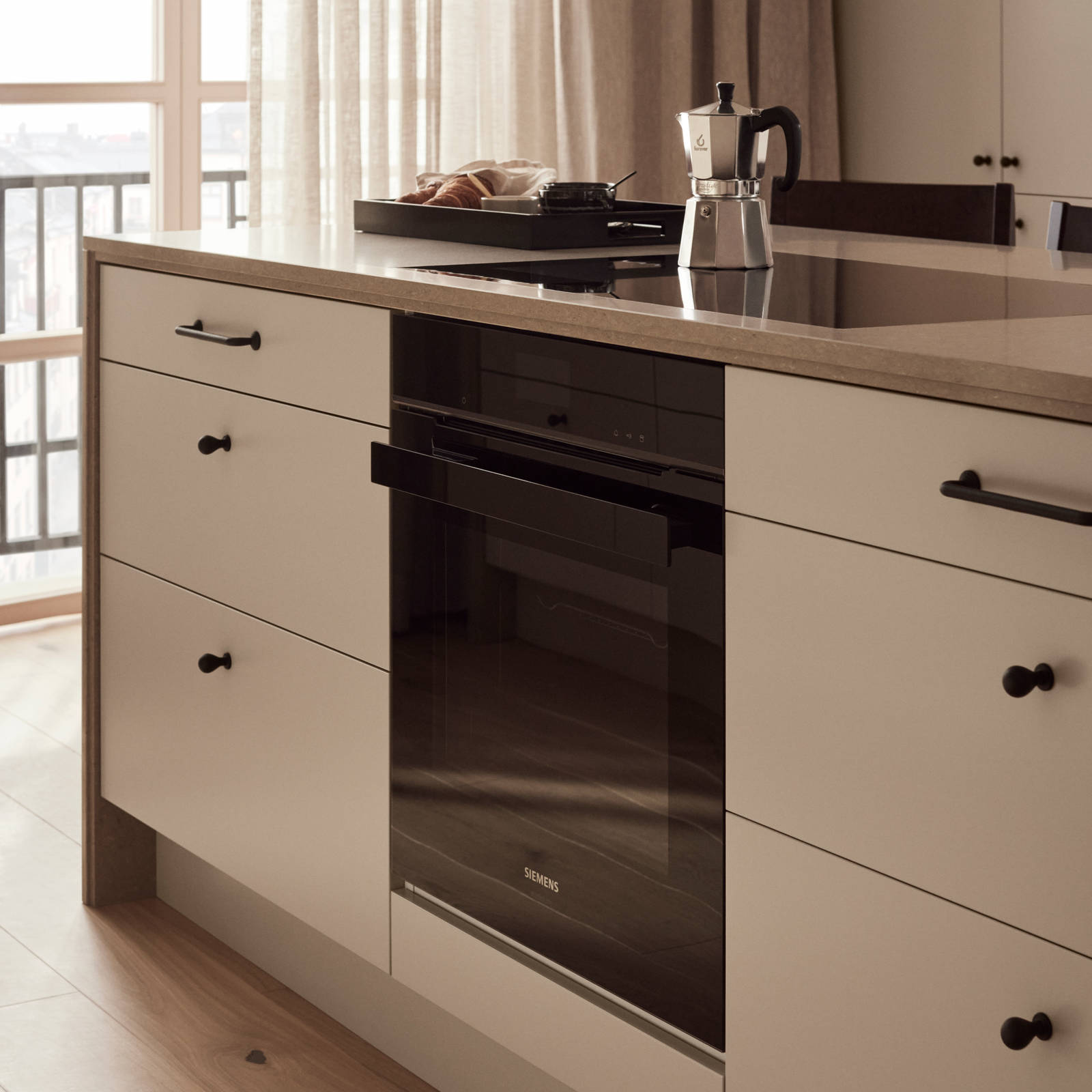 Inbyggnadsugn och induktionshäll från Siemens i köksö i köket med beige slät kökslucka Arkitekt Plus gryning, bänkskiva och dekorativ kant i kompositsten, svarta knoppar