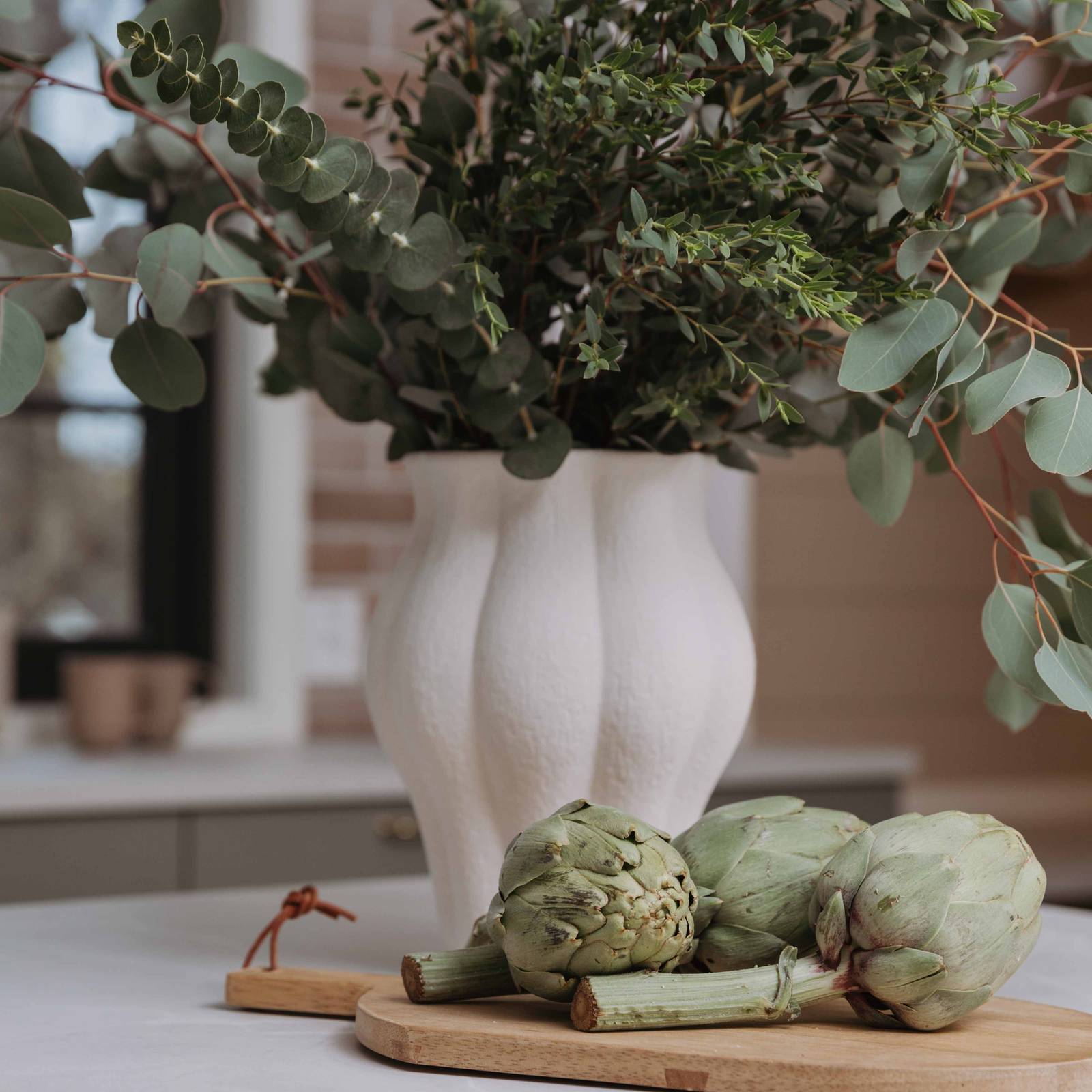 En keramikkvase med grønne planter og et skjærebrett i tre med ertebelger på kjøkkenbenken i Stine Hartmanns Uno Furu kjøkken fra Sigdal