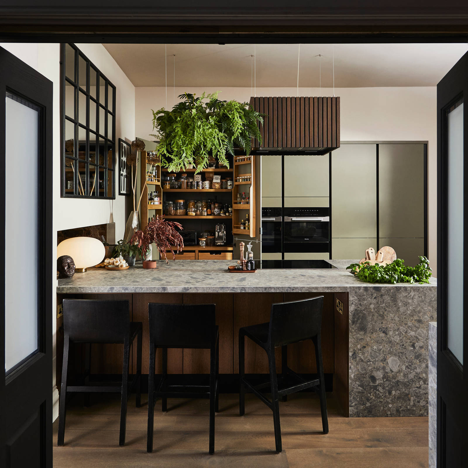 View through opened dark wood doors of the biophilic Dramatic neutral Balmoral Soho and Nordic Nature kitchen from Magnet, with the pantry larder cabinet opened revealing oak interiors.