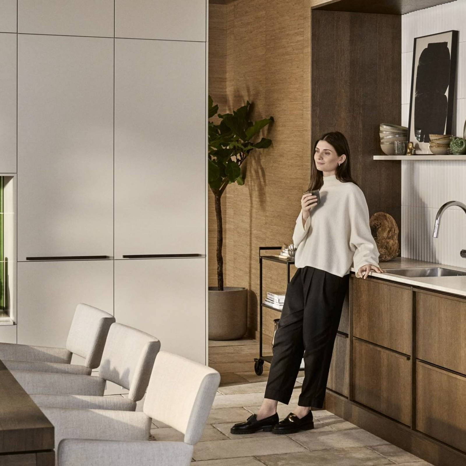 Kvinde læner sig mod mørk eg Forever Oak køkkenbænken fra INVITA i køkkenet med overskabe og højskabe i Athena Sand