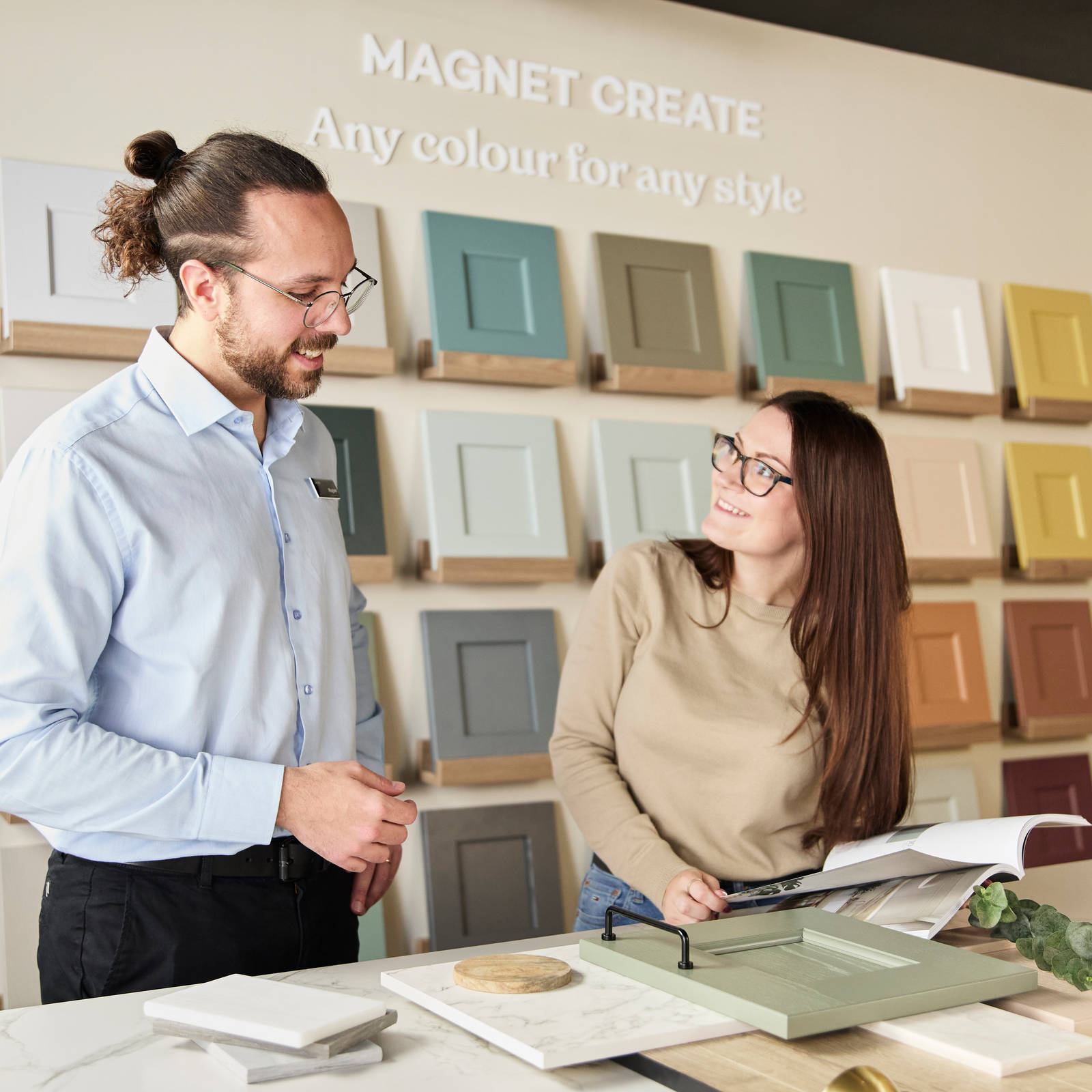 Kitchen designer showing samples of fronts, worktops and handles to a woman in a Magnet Showroom and referencing a brochure.