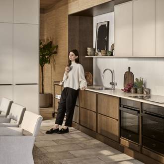 Kvinde læner sig mod mørk eg Forever Oak køkkenbænken fra INVITA i køkkenet med overskabe og højskabe i Athena Sand