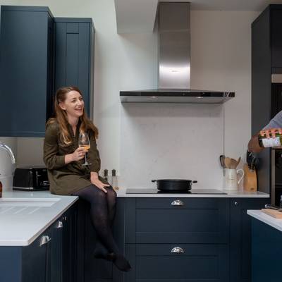 Amy sitting on the kitchen counter with a wine glass in hand while Kamal stands by the kitchen island pouring a glass of wine for himself.