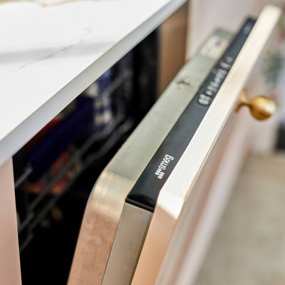 Close-up view of slightly open integrated Smeg dishwasher in Ludlow Chalk Blush cabinetry from Magnet, with a brass handle and Dekton worktop.