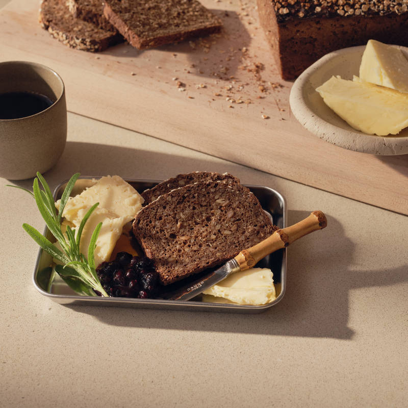 Et skjærebrett med brød, en kniv og en tallerken med en sandwich på den grå kjøkkenbenken i eikekjøkkenet Favn Natur fra Sigdal i eikefiner med vertikal trestruktur.