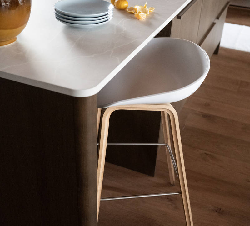 Keittiösaareke pyöristetyllä kulmalla, kaapit laatikoilla ja istumatilaa valkoisella ja puisella baarijakkaralla