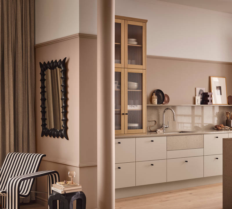 Design-fåtölj, bord, pelare och i bakgrunden beige köket Arkitekt plus gryning från marbodal med vitrinskåp med träram i natur ek och räfflat glas