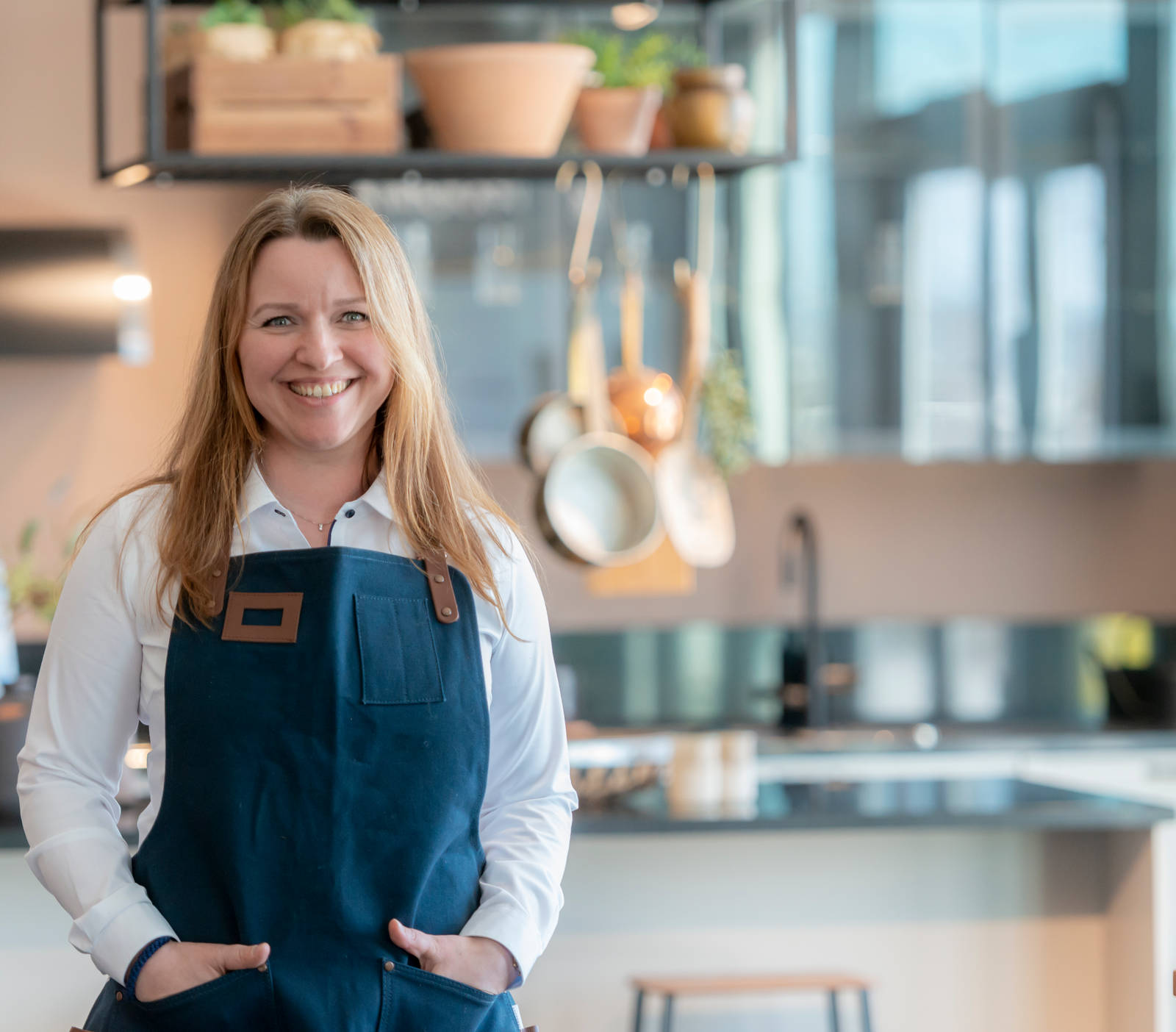 Kjøkkendesigner i lys grå skjorte og blått forkle smiler til kamera utenfor Sigdals butikk