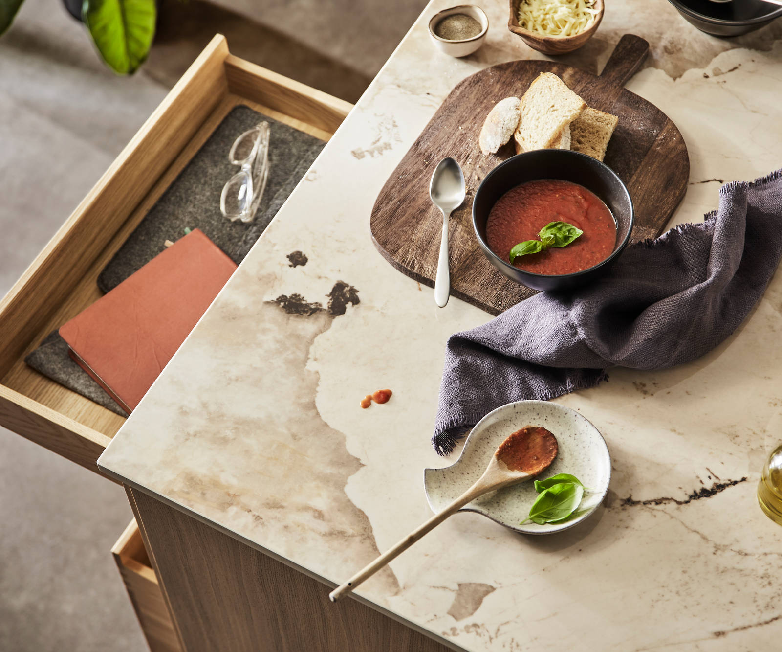 Bird view of kitchen island with Dekton Khalo Worktop, ceramic bowls containing tomato soup, bread and olive oil while an oak drawer is opened.
