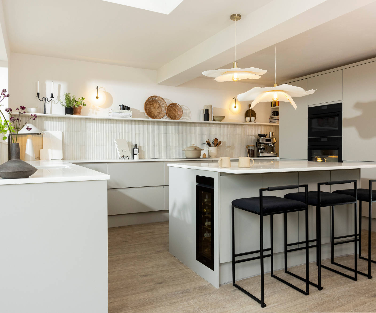 Angled view of kitchen island in Yasmin Wyatt's neutral shade Luna kitchen from Magnet with a white Corian worktop, integrated whitegoods and black metal chairs by the kitchen island bar area.