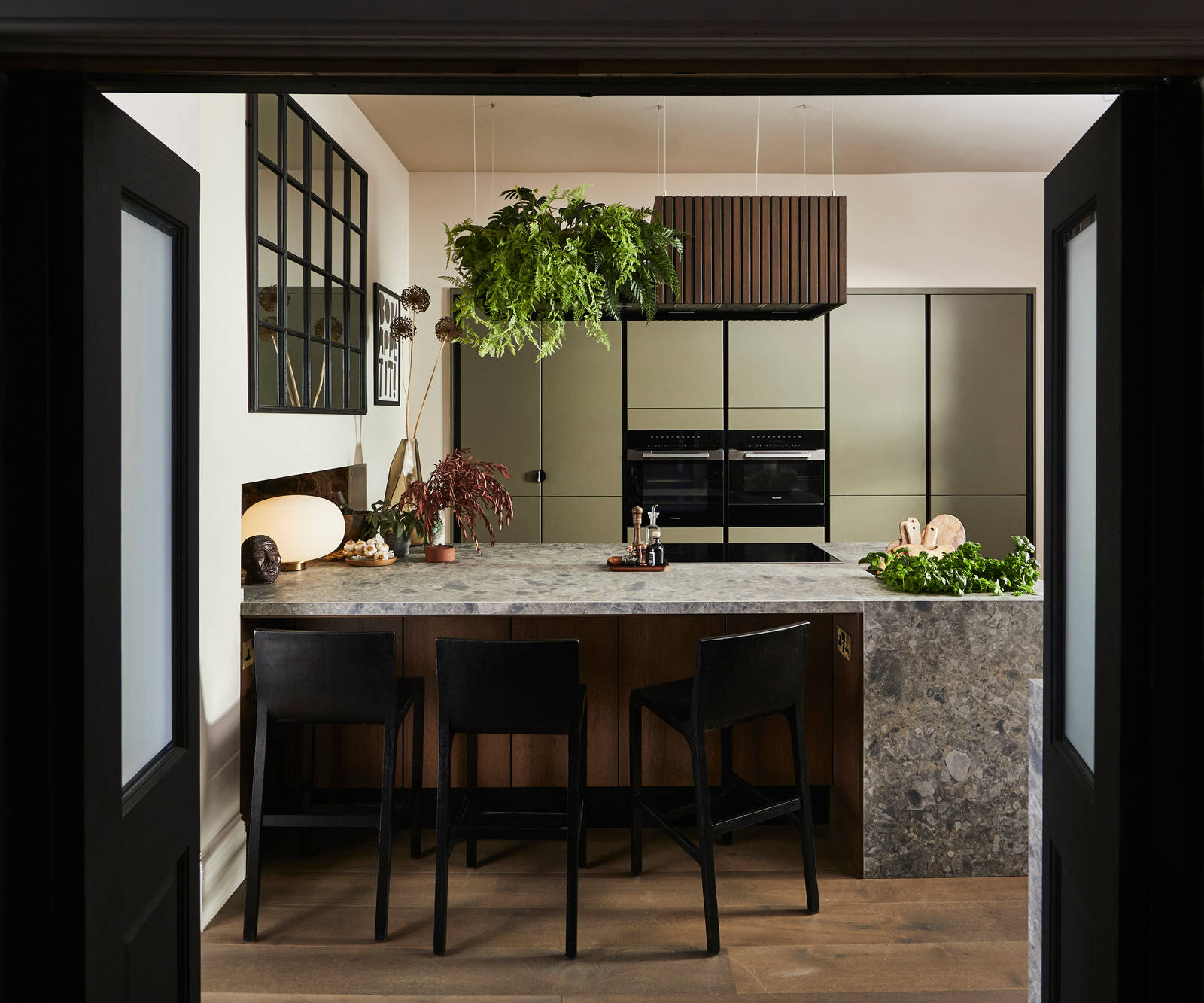 View through opened dark wood doors of the biophilic Dramatic neutral Balmoral Soho and Nordic Nature kitchen from Magnet and matching dark chairs by the bar area of the kitchen island.