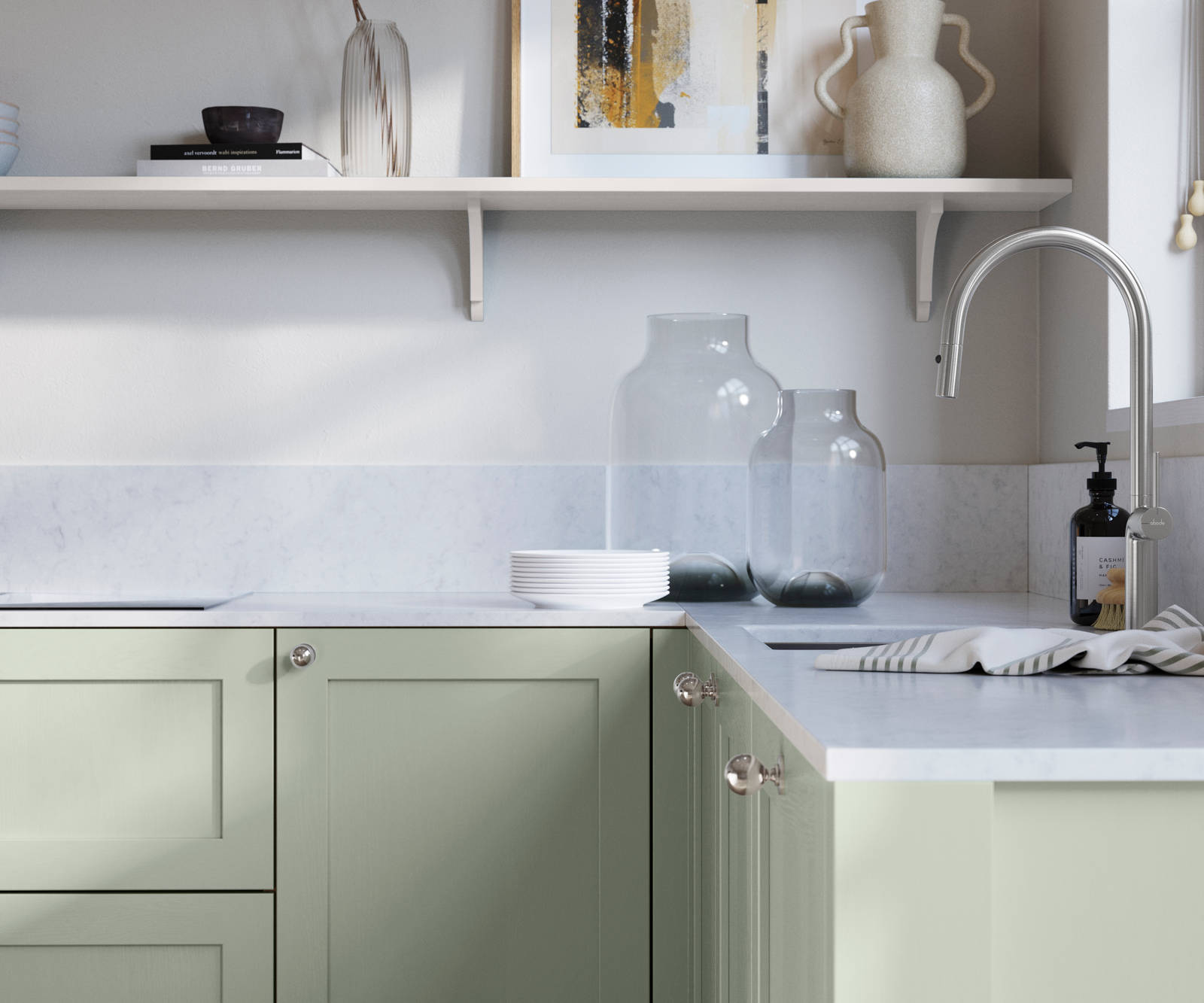 Wardley Seagrass kitchen cabinetry with steel knobs, steel tap, white marbled worktop and matching backsplash. An open shelf on the wall above cabinets with ceramics and a vase of flowers.