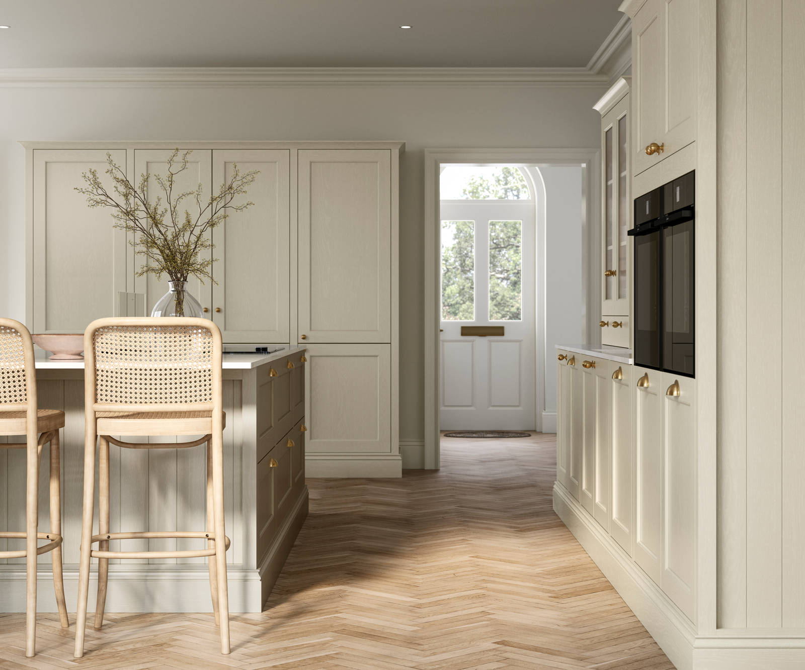 Traditional Shaker-style kitchen Ludlow Parchment with tactile woodgrain, tall cabinets, brass cup handles and knobs, integrated ovens, a kitchen island with rattan bar stools, a vase holding foliage, and light wood herringbone flooring.
