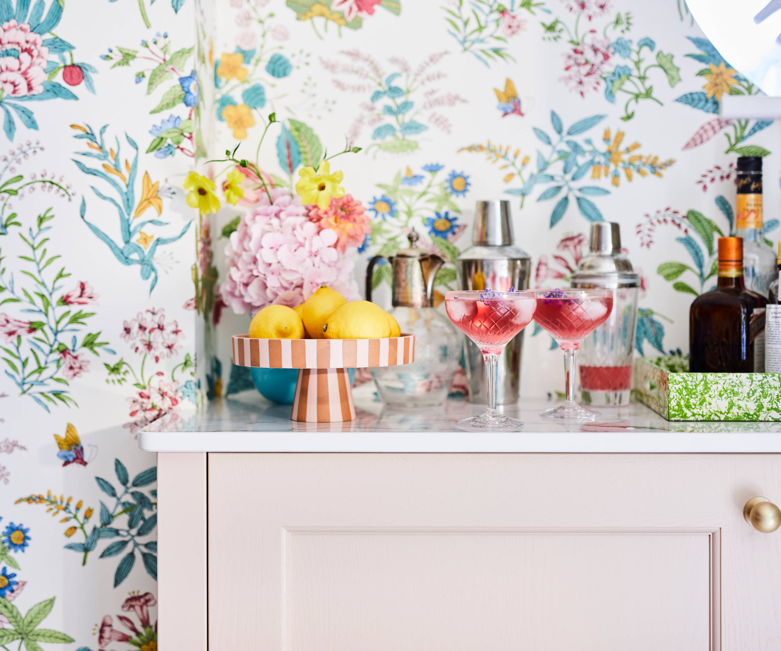 Cocktail corner in Sophie Robinsons maximalist Ludlow kitchen from Magnet, with two pink drinks poured in glasses, floral pattern wallpaper.
