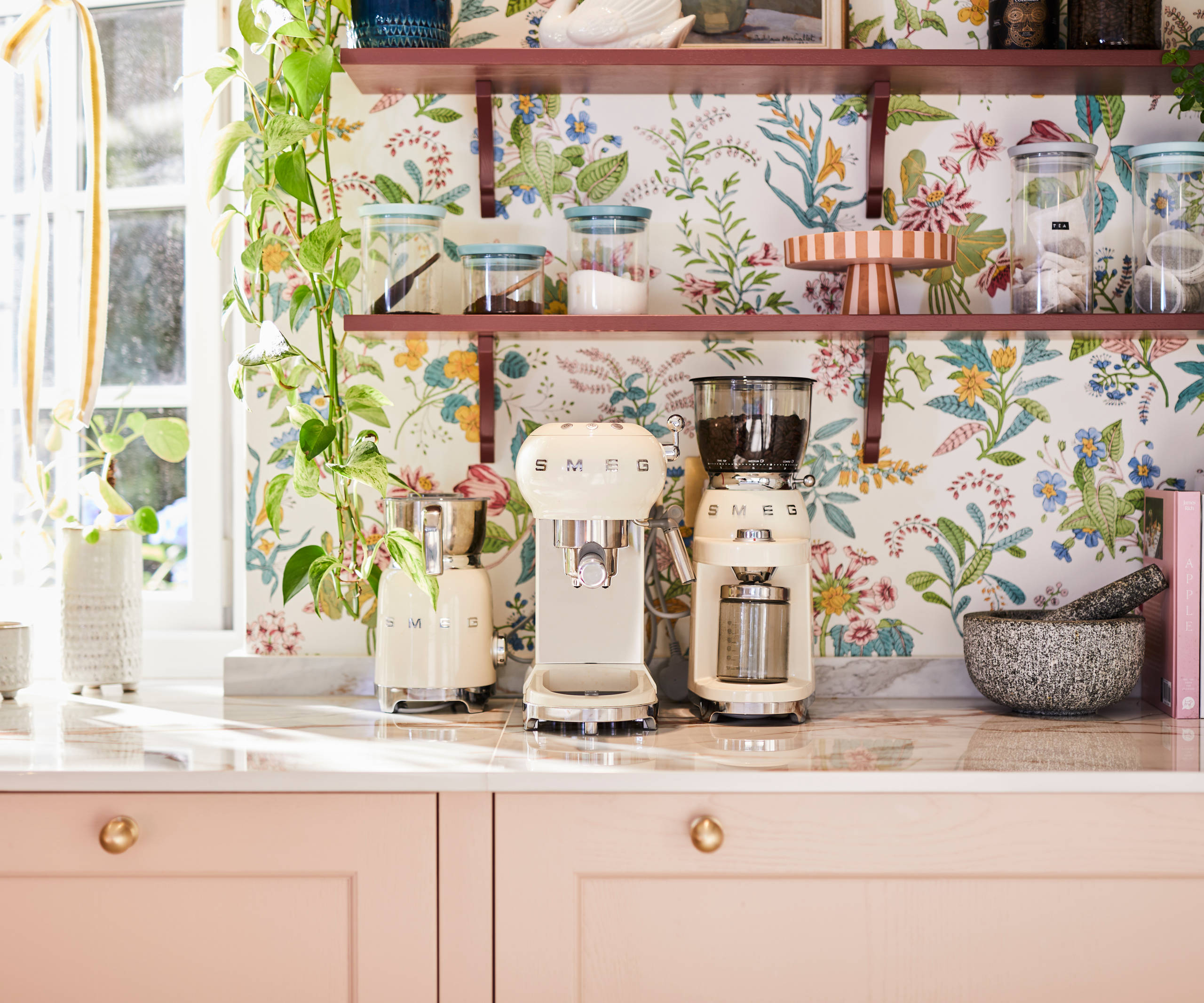 Close-up of Smeg coffee machines on counter top in the coffee area in Sophie Robinson's maximalist kitchen, with shaker style Ludlow Chalk blush cabinetry, open shelving on a wall with a floral wallpaper.