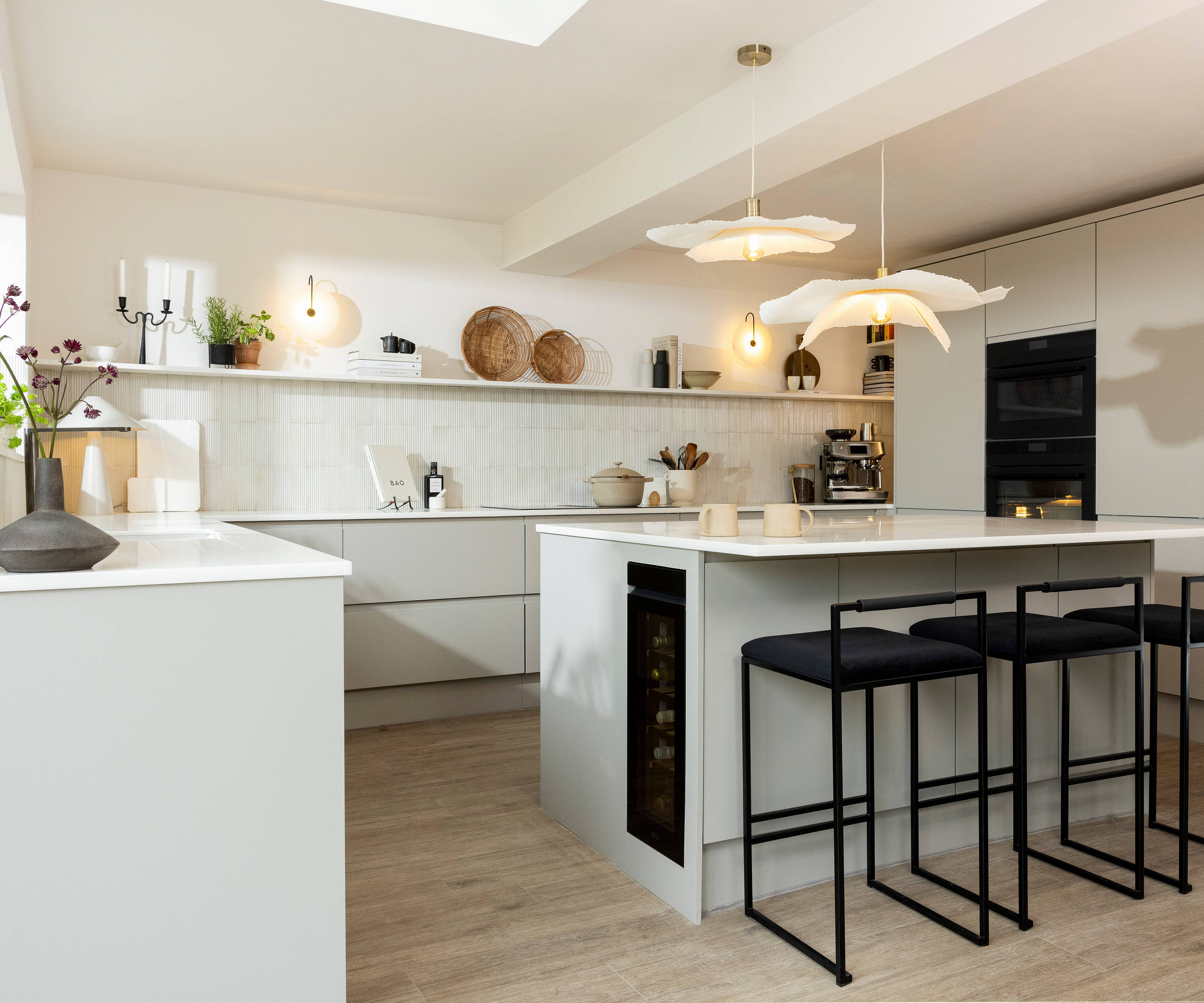 Angled view of kitchen island in Yasmin Wyatt's neutral shade Luna kitchen from Magnet with a white Corian worktop, integrated whitegoods and black metal chairs by the kitchen island bar area.