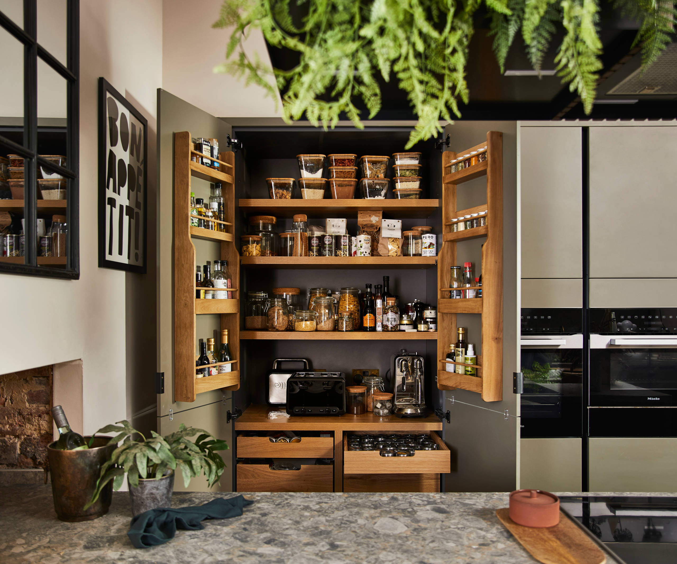 View from kitchen island of opened pantry larder with oak interior full of glass jars containing dry goods, a toaster and spices
