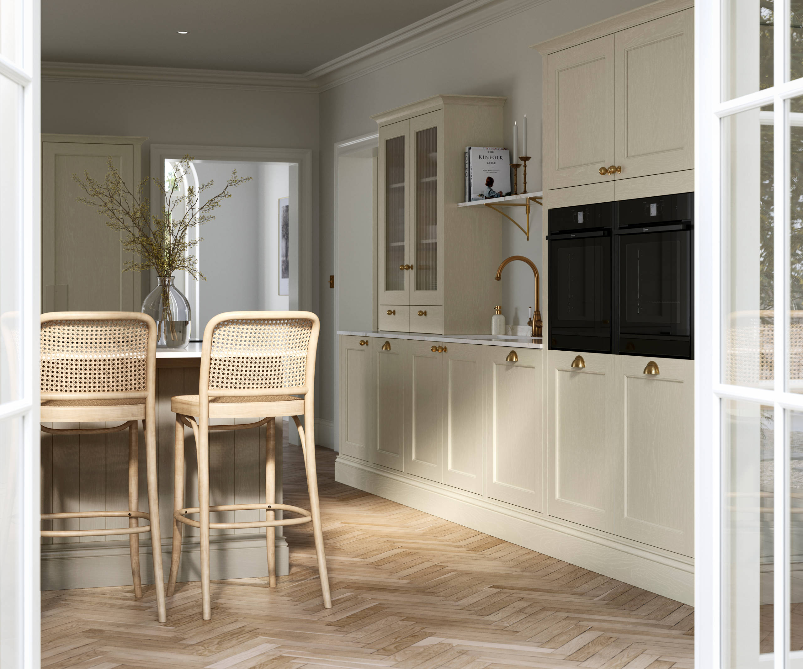 View from patio through opened white frame glass doors of traditional Shaker-style kitchen Ludlow Parchment with tactile woodgrain, tall cabinets, brass cup handles and knobs, integrated ovens, a kitchen island with rattan bar stools, a vase holding foliage, and light wood herringbone flooring.
