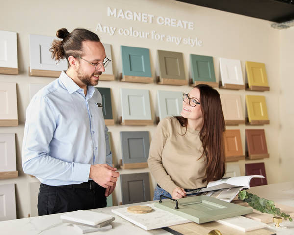 Kitchen designer showing samples of fronts, worktops and handles to a woman in a Magnet Showroom and referencing a brochure.