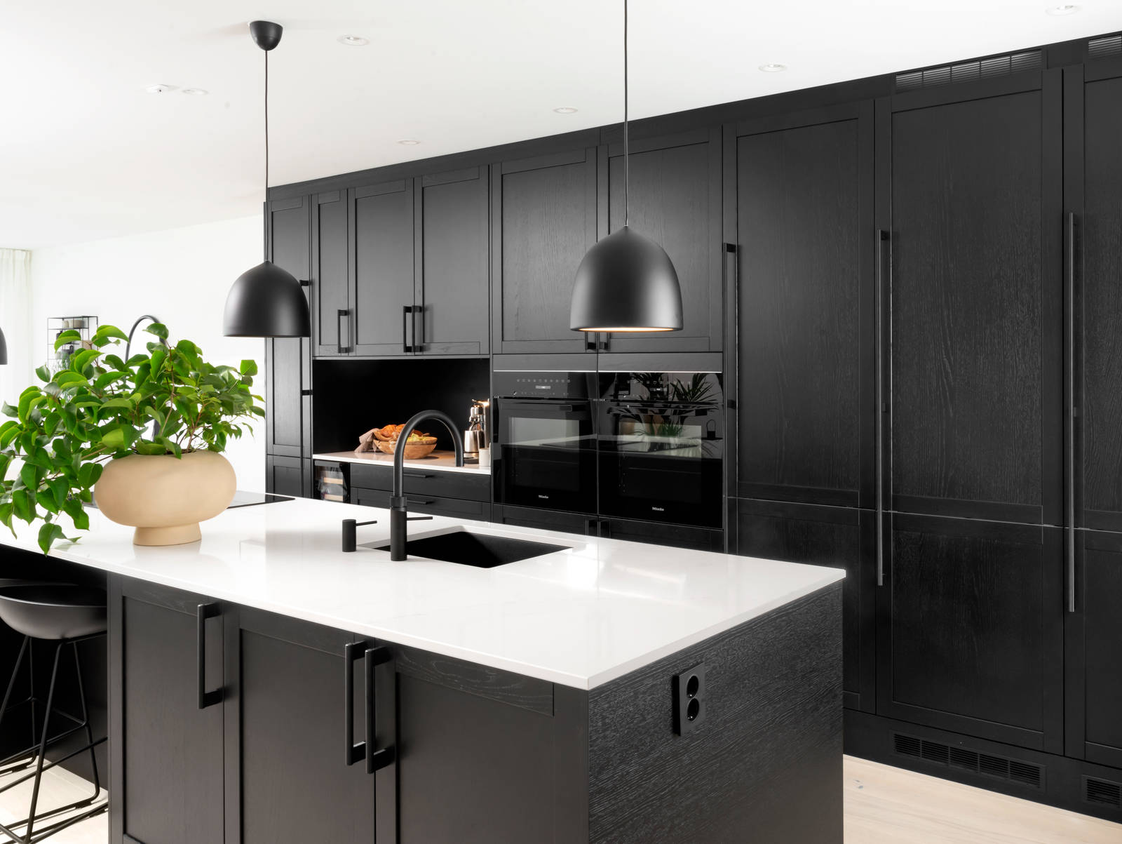 Et nydelig svart kjøkken i lun eik med kjøkkenøy dekket av lys komposittstein og stilige matt-svarte håndtak.