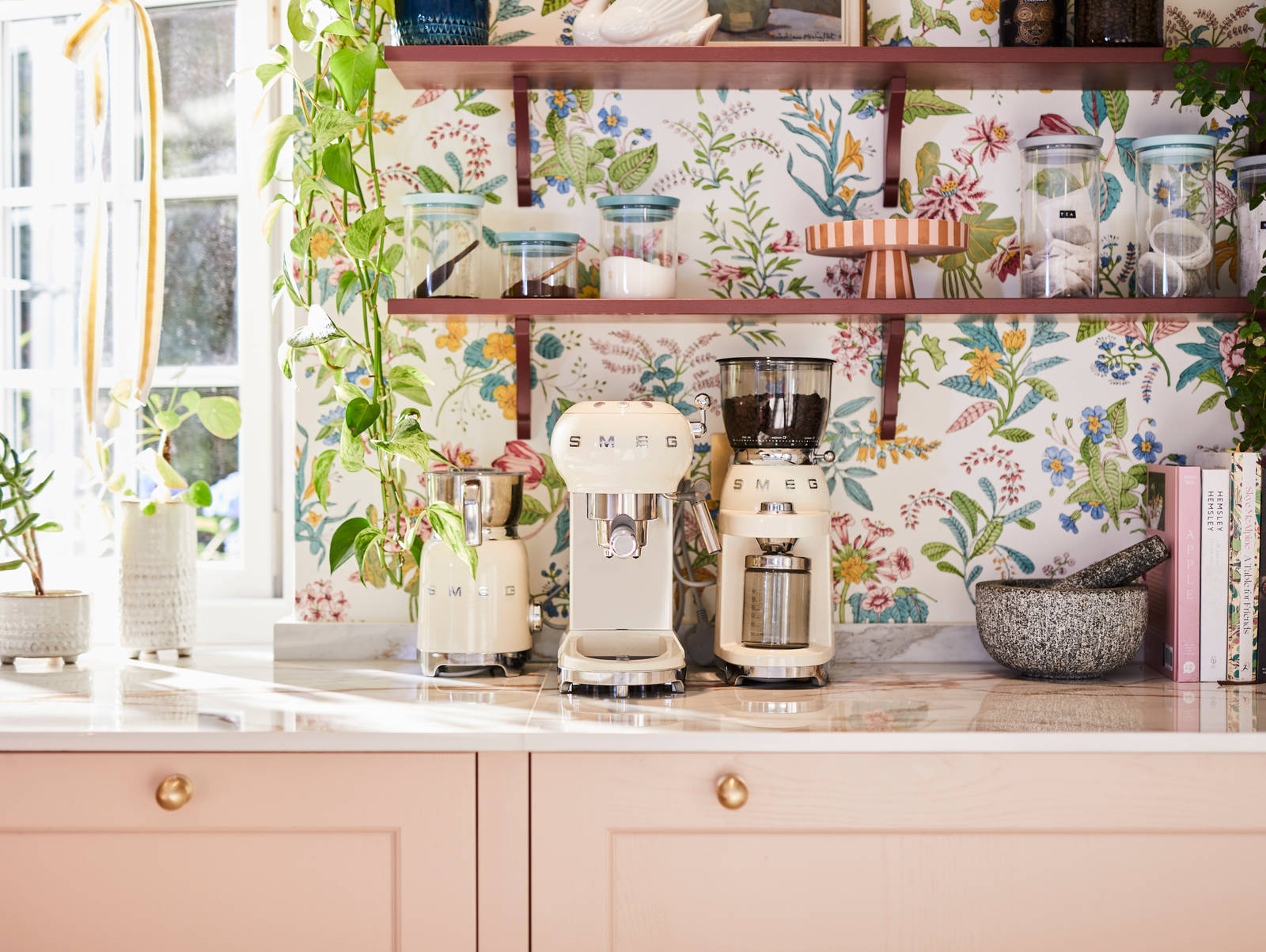Close-up of Smeg coffee machines on counter top in the coffee area in Sophie Robinson's maximalist kitchen, with shaker style Ludlow Chalk blush cabinetry, open shelving on a wall with a floral wallpaper.