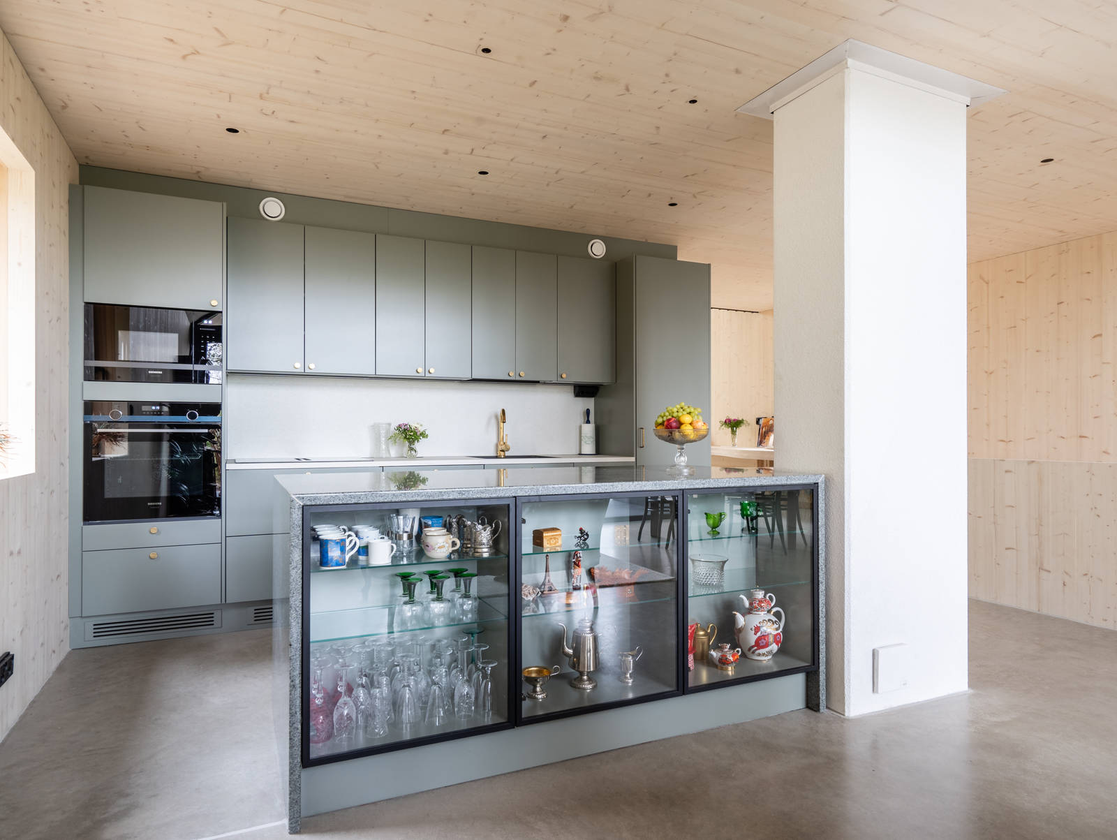 Vihreä keittiö Novartin luonnonvärisessä Havu-sävyssä, messinkinuppeilla, graniittitasoilla ja lasiovilla, joissa on alumiinikehykset keittiön altaassa.