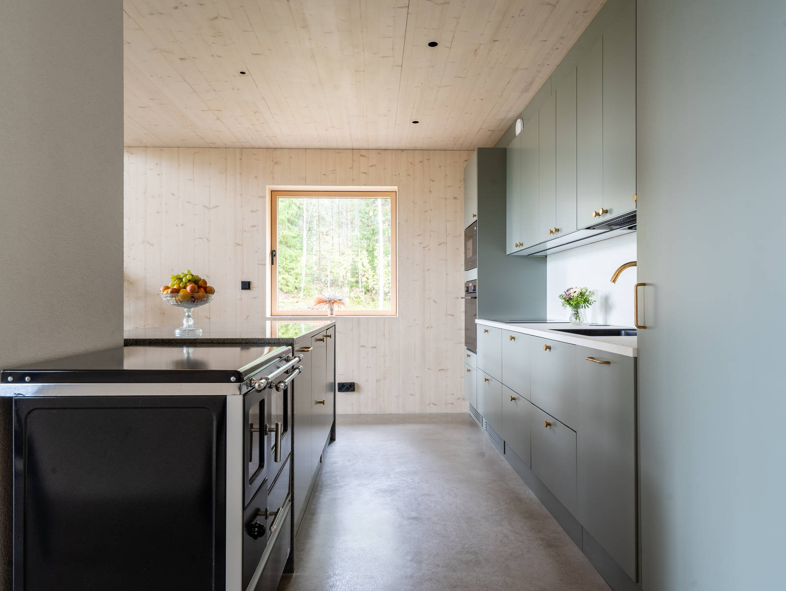 Vihreä keittiö Novartin luonnonvärisessä Havu-sävyssä, messinkinuppeilla, graniittitasoilla ja lasiovilla, joissa on alumiinikehykset keittiön altaassa.