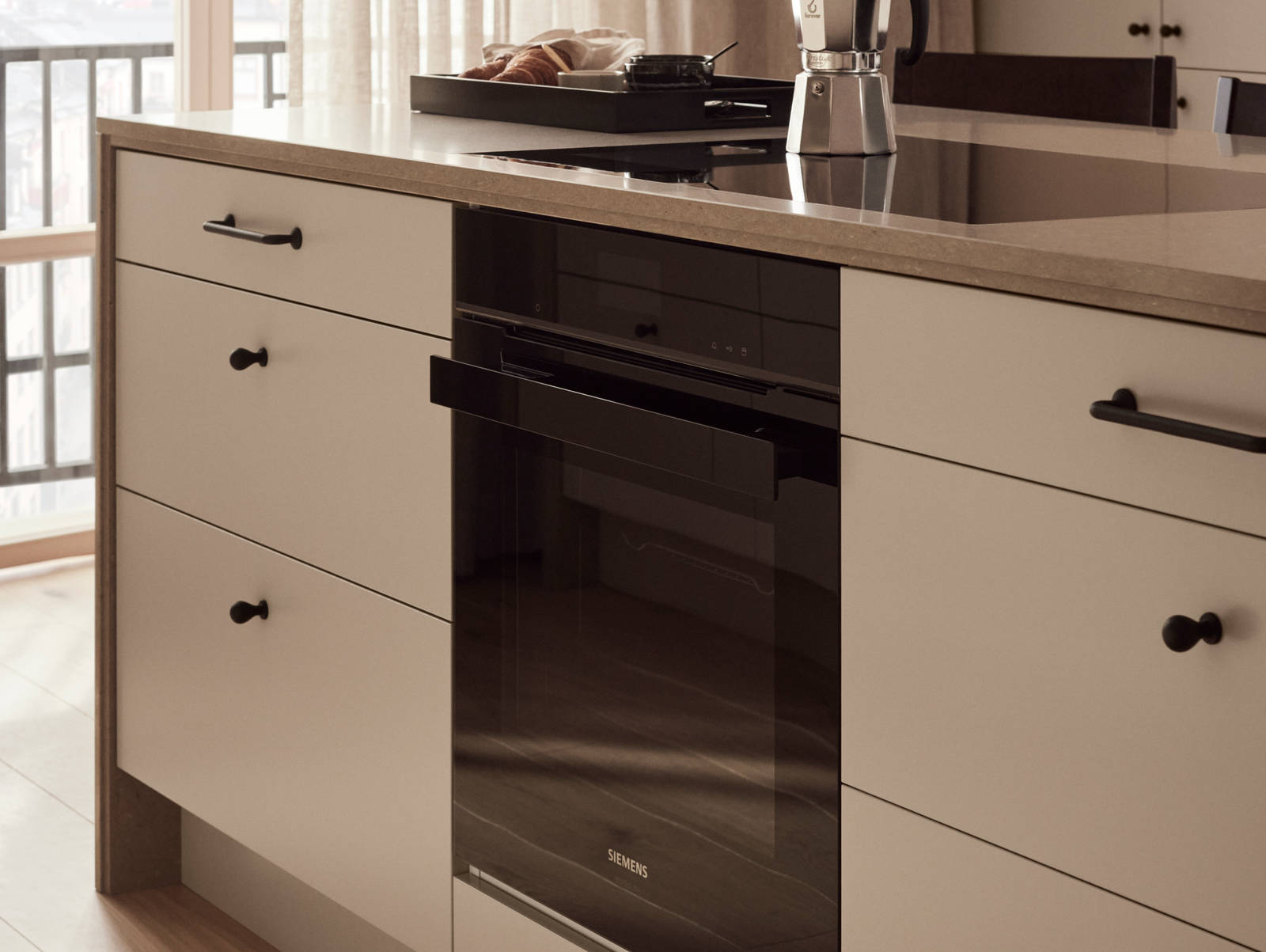 Inbyggnadsugn och induktionshäll från Siemens i köksö i köket med beige slät kökslucka Arkitekt Plus gryning, bänkskiva och dekorativ kant i kompositsten, svarta knoppar