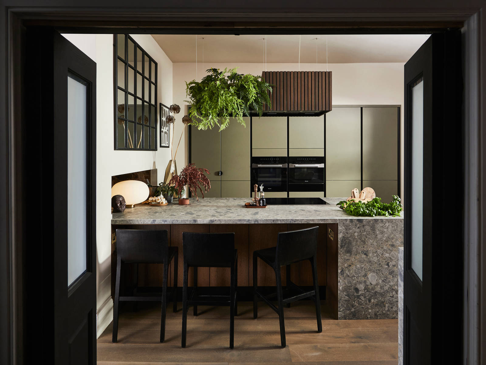 View through opened dark wood doors of the biophilic Dramatic neutral Balmoral Soho and Nordic Nature kitchen from Magnet and matching dark chairs by the bar area of the kitchen island.