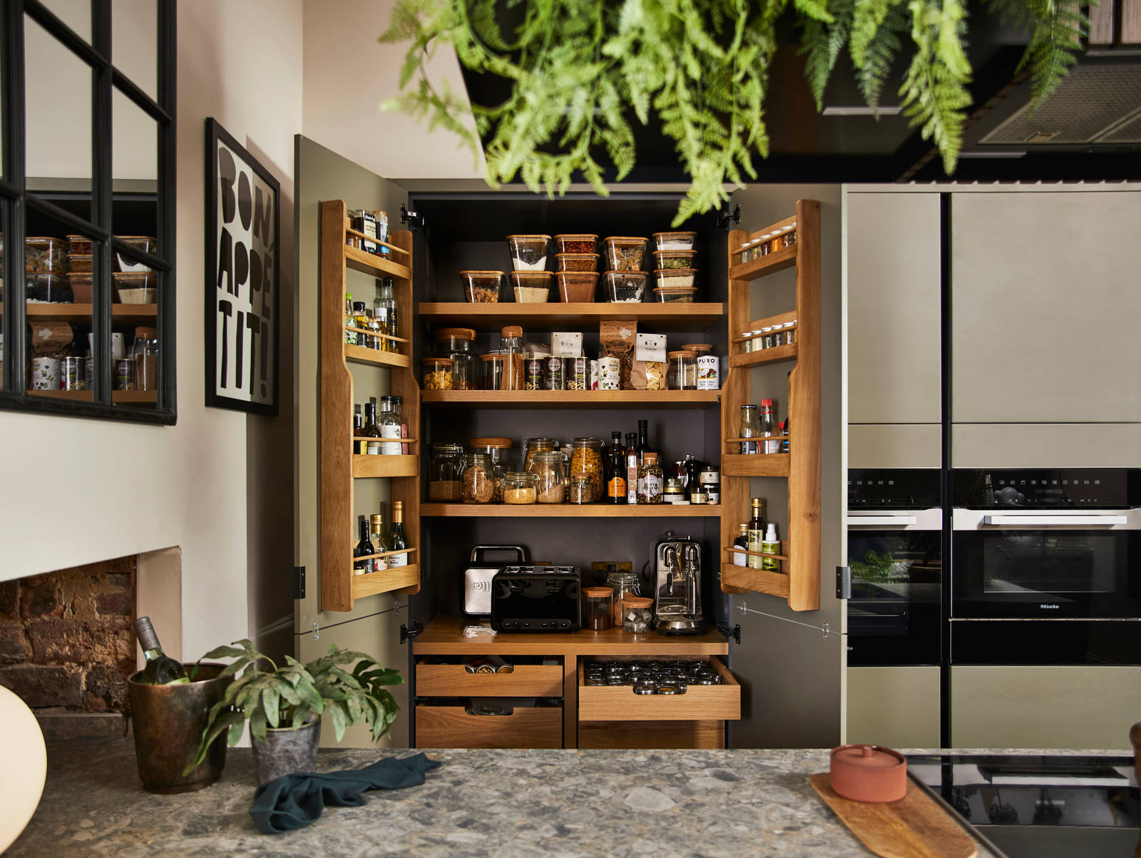 View from kitchen island of opened pantry larder with oak interior full of glass jars containing dry goods, a toaster and spices