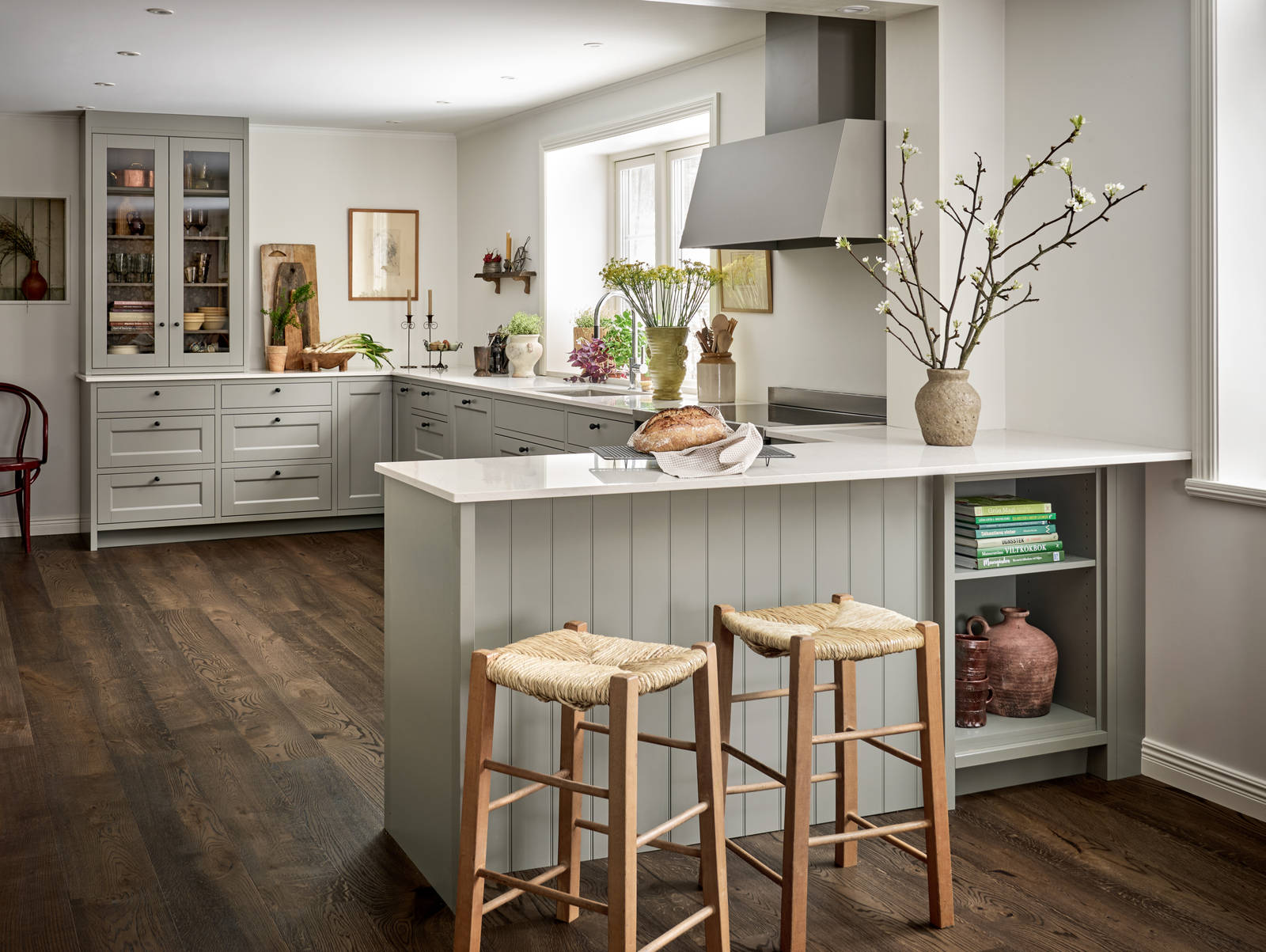 Malin Berghagens kök från Marbodal, lantliga Fagerö i kulören Frö med vit och grå marmorerad bänkskiva, med halvt fristående köksö med två barstolar och en öppen hylla för böcker och keramik.