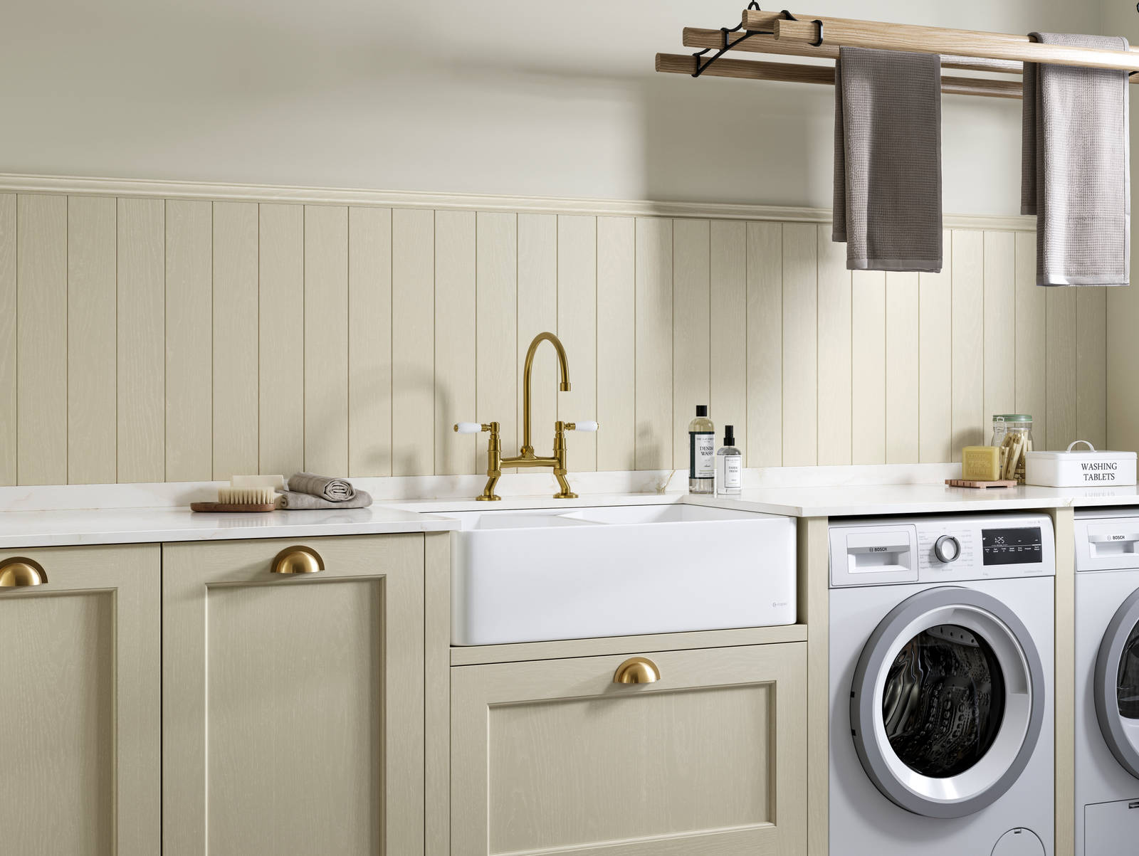 Traditional Shaker-style Ludlow Parchment and French Clay utility room with white ceramic sink, washing machine and tumble dryer with white marbled Silestone worktop and upstand, wallpanel matching the cabinetry, traditional brass tap and towels hanging on wood drying rack.