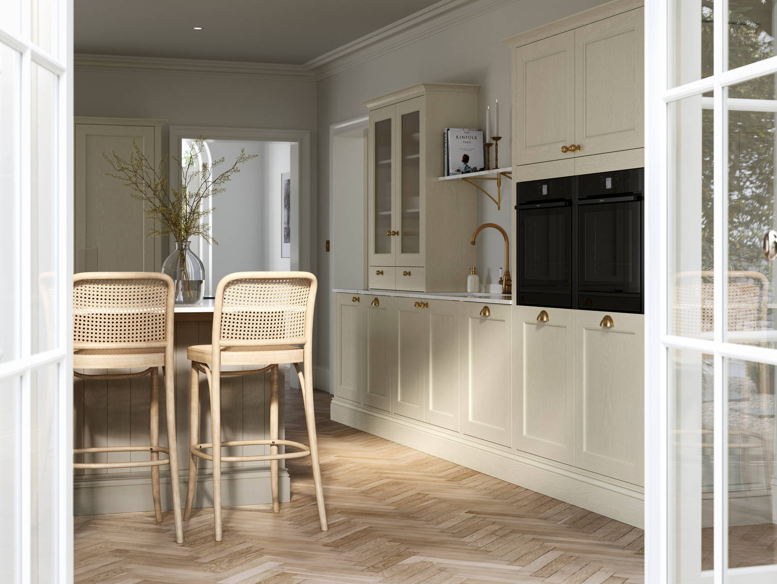 View from patio through opened white frame glass doors of traditional Shaker-style kitchen Ludlow Parchment with tactile woodgrain, tall cabinets, brass cup handles and knobs, integrated ovens, a kitchen island with rattan bar stools, a vase holding foliage, and light wood herringbone flooring.