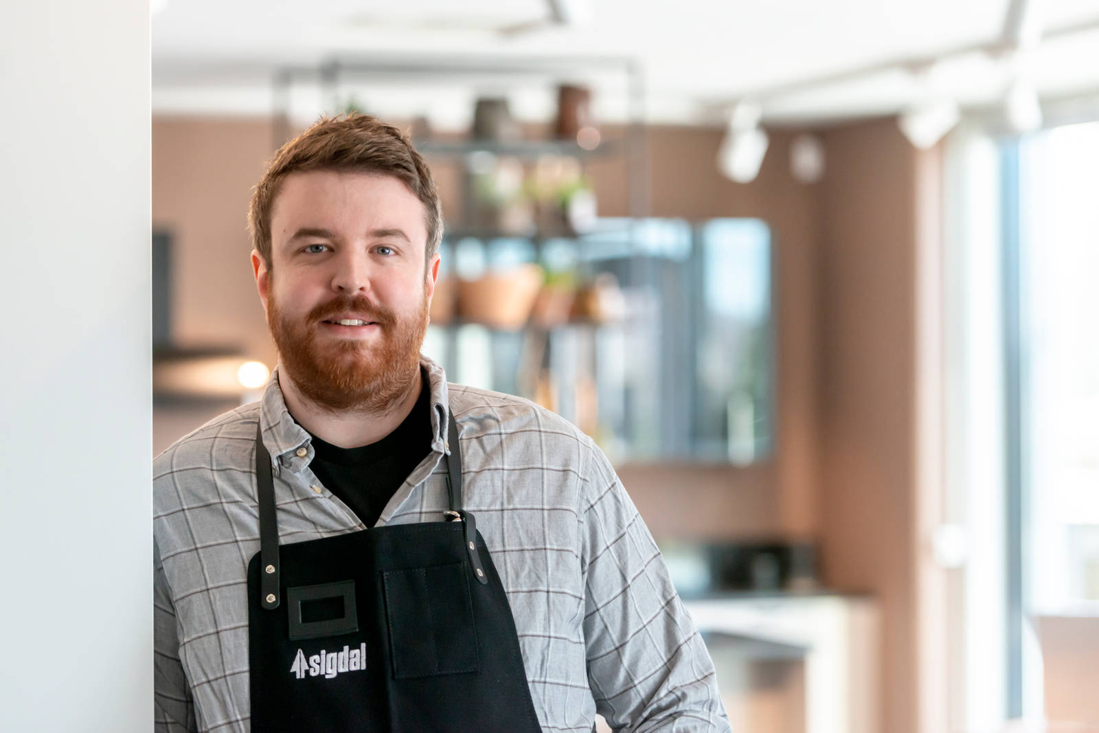 Kjøkkendesigner i lys grå skjorte og blått forkle smiler til kamera utenfor Sigdals butikk