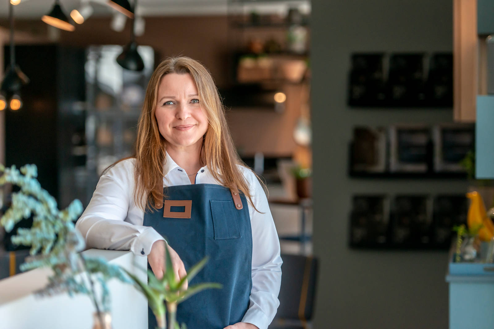 Kjøkkendesigner i lys grå skjorte og blått forkle smiler til kamera utenfor Sigdals butikk