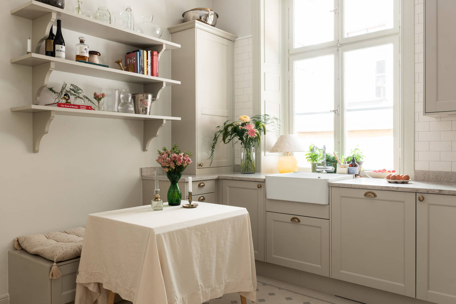 Sandra Beijers kök från Marbodal, lantliga luckan Fagerö i beige kulören Sand, med bronsfärgade handtag, diskho i vitt porslin, öppna vägghyllor i matchande beige kulör, en vit duk och mässingsljusstakar på matbordet.