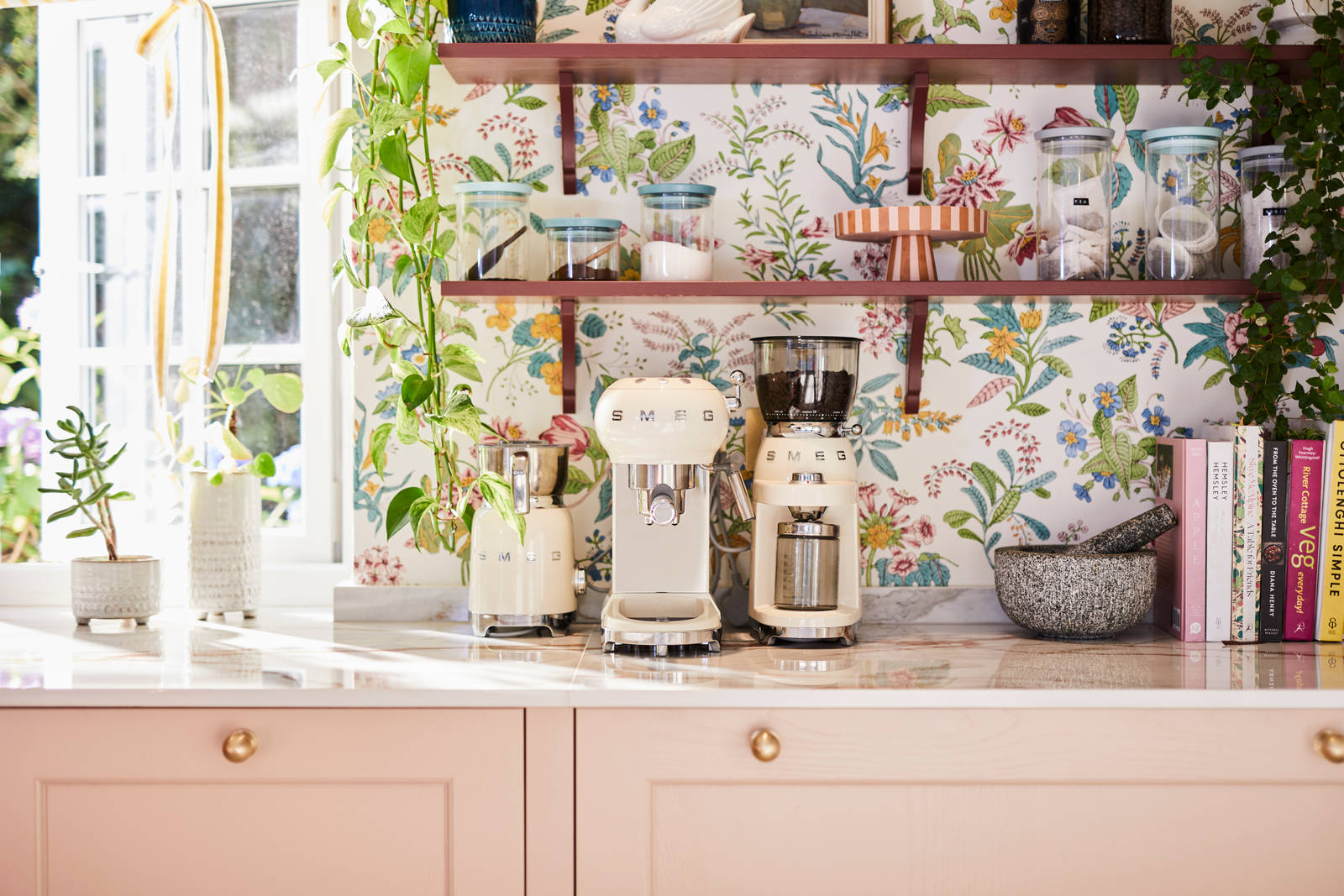 Close-up of Smeg coffee machines on counter top in the coffee area in Sophie Robinson's maximalist kitchen, with shaker style Ludlow Chalk blush cabinetry, open shelving on a wall with a floral wallpaper.