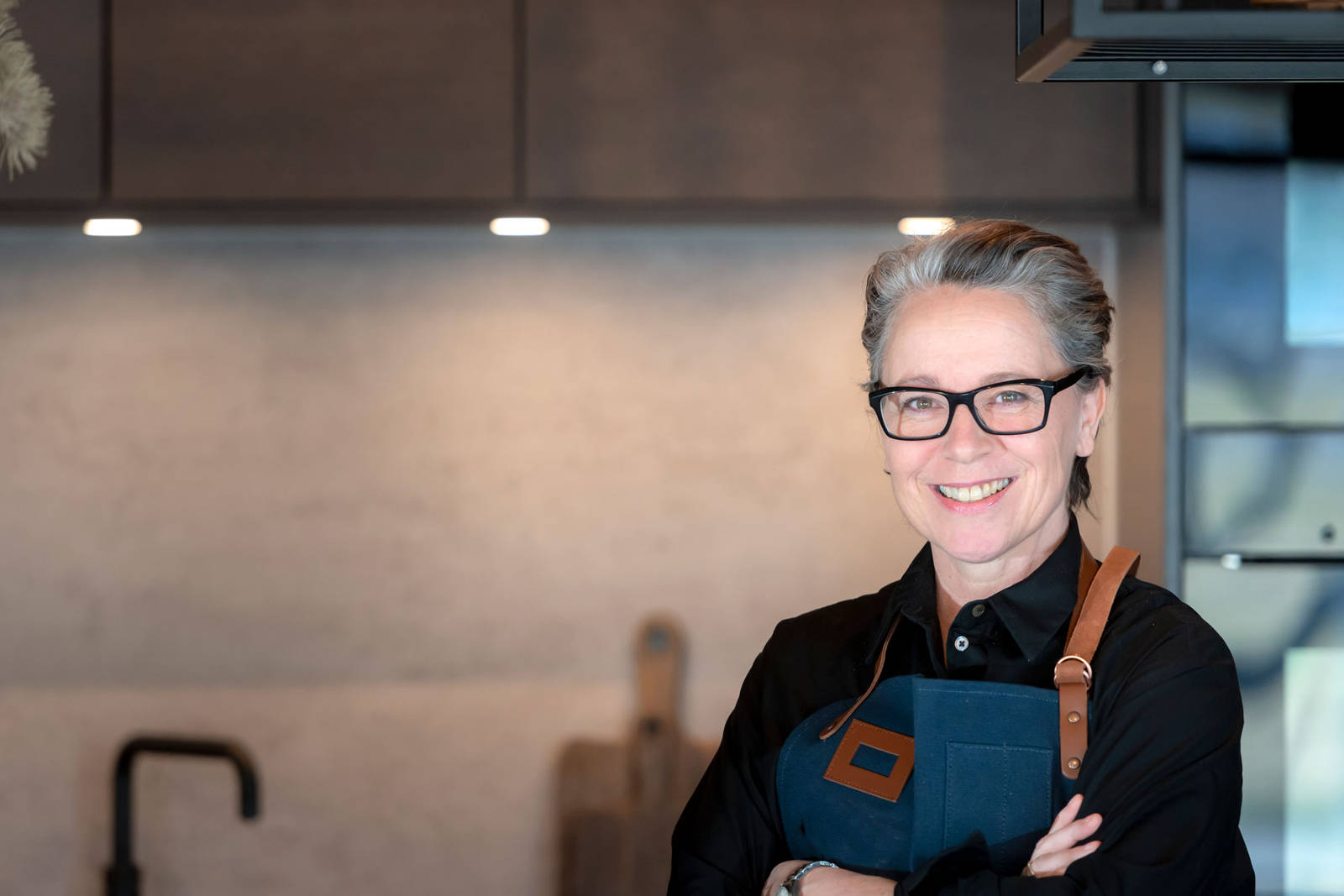 Kjøkkendesigner med svart skjorte og blått forkle smiler til kamera i Sigdals butikk