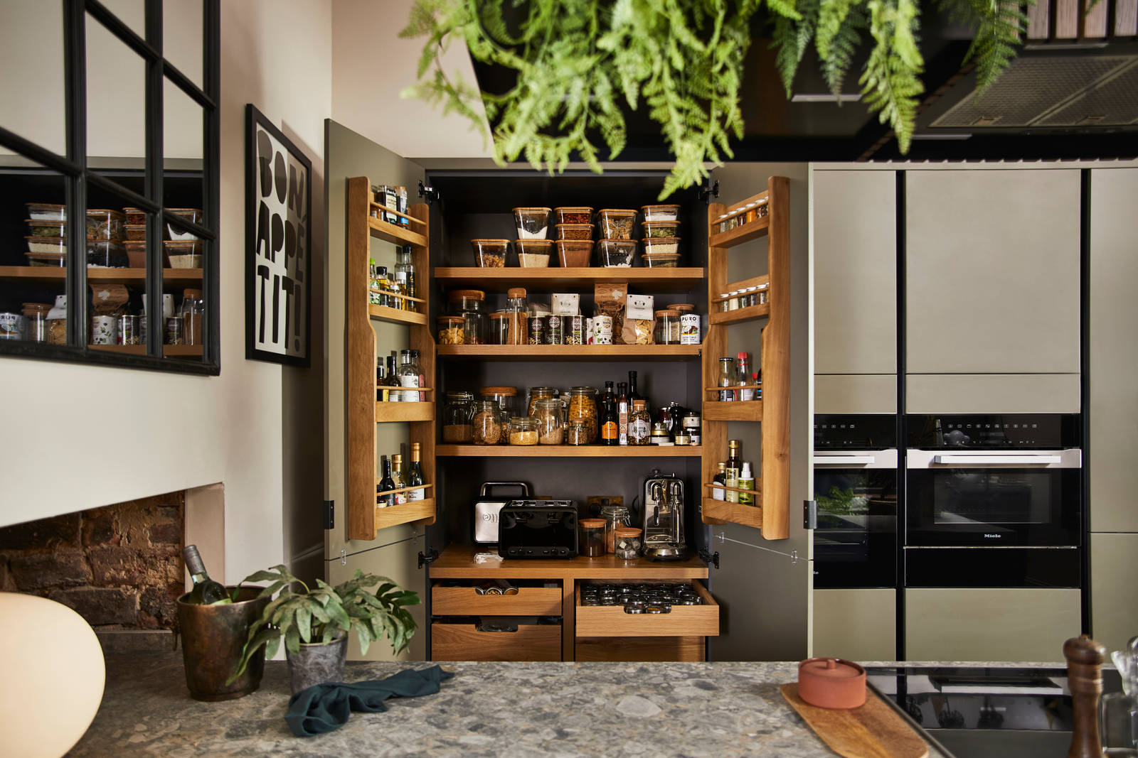 View from kitchen island of opened pantry larder with oak interior full of glass jars containing dry goods, a toaster and spices
