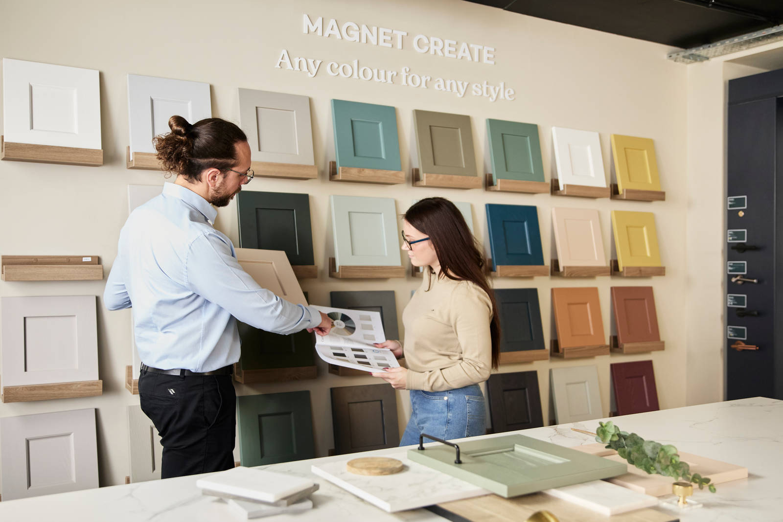 Kitchen designer holding a colour sample while pointing in the brochure held by a customer in a Magnet Showroom.
