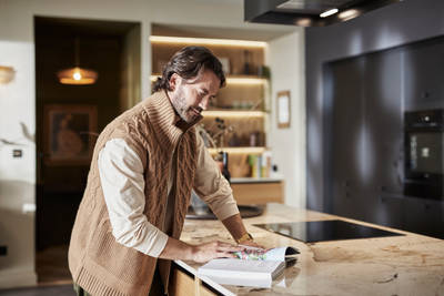 Man wearing clothing in earth tones reading a cook book at the kitchen island counter in Hoxton Sumi Black and Nordic Craft kitchen from Magnet.