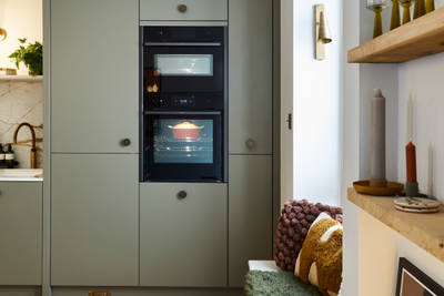 Soho Balmoral tall cabinets with integrated ovens and a bench in the window, next to a wall of open wood shelves decorated with plants, candles and wine glasses.