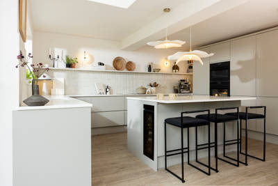Angled view of kitchen island in Yasmin Wyatt's neutral shade Luna kitchen from Magnet with a white Corian worktop, integrated whitegoods and black metal chairs by the kitchen island bar area.