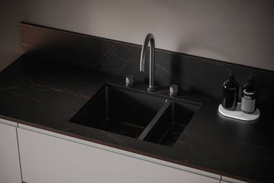 The J-pull handle of Magnet's Duxbury kitchen range is seen with a dark kitchen worktop and modern integrated double sink and arched tap.
