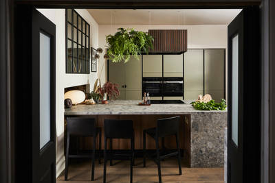 View through opened dark wood doors of the biophilic Dramatic neutral Balmoral Soho and Nordic Nature kitchen from Magnet, matching dark wood chairs by kitchen island bar seating area.