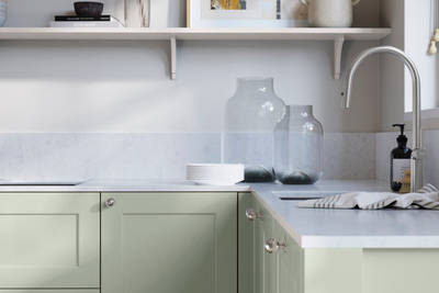 Ludlow, a traditional Shaker-style premium feel kitchen range with a visible woodgrain on its panelled doors here shown in the light shade Seagrass styled with steel knobs, steel tap, a white marbled worktop with matching splashback.