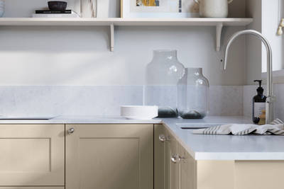 Ludlow, a traditional Shaker-style premium feel kitchen range with a visible woodgrain on its panelled doors here shown in the neutral shade Pebble path styled with steel knobs, steel tap, a white marbled worktop with matching splashback.